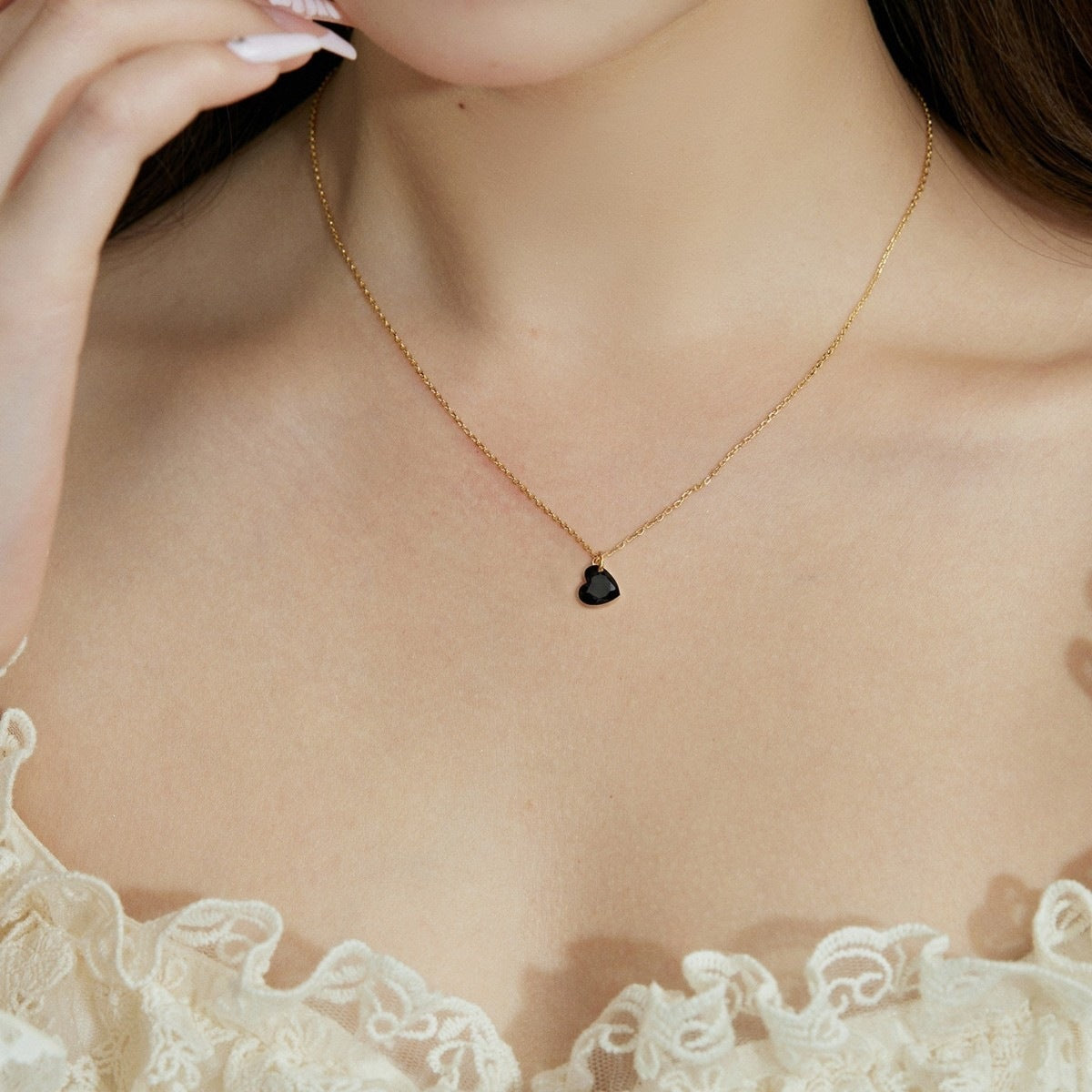 A delicate gold chain necklace with a small black heart-shaped pendant, worn by a model in a close-up shot.