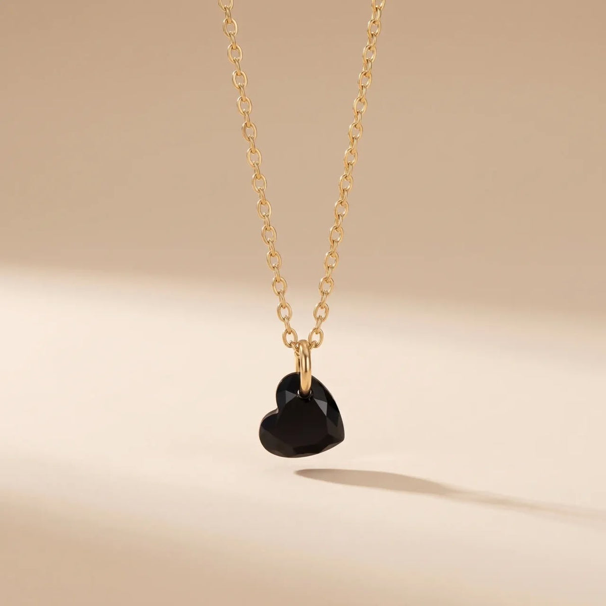 A gold chain necklace with a faceted black heart pendant, suspended against a warm beige background with a soft shadow.