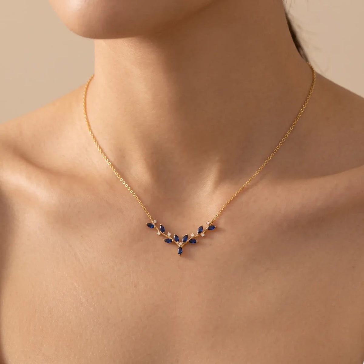A delicate gold chain necklace with a small V-shaped pendant featuring dark blue teardrop stones and tiny clear accent stones, shown worn on a model's neck against a warm beige background.