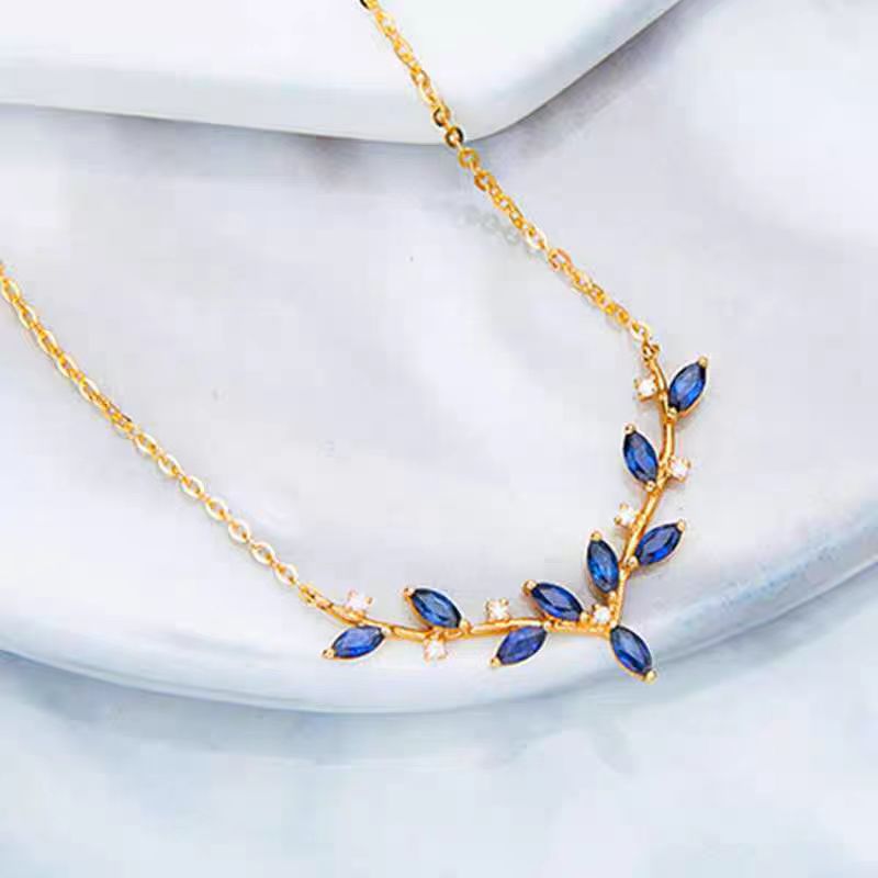 Gold leaf-shaped necklace with blue marquise and oval gemstones, accented by small clear stones, displayed on a white marble surface.