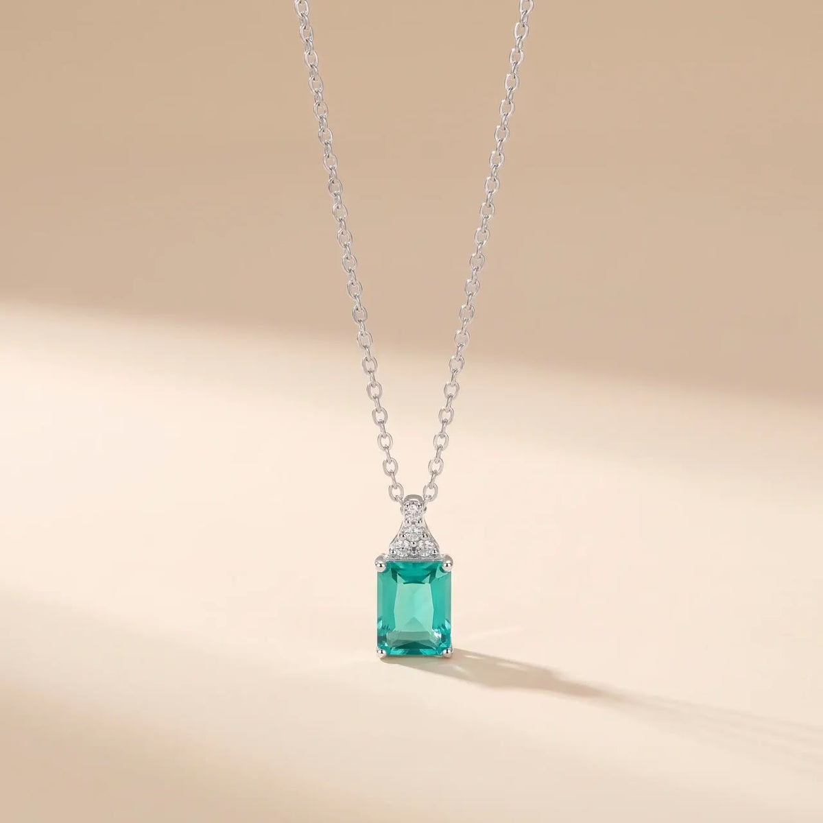A silver necklace with a rectangular emerald-cut green gemstone pendant and small clear accent stones, shown on a warm beige background.