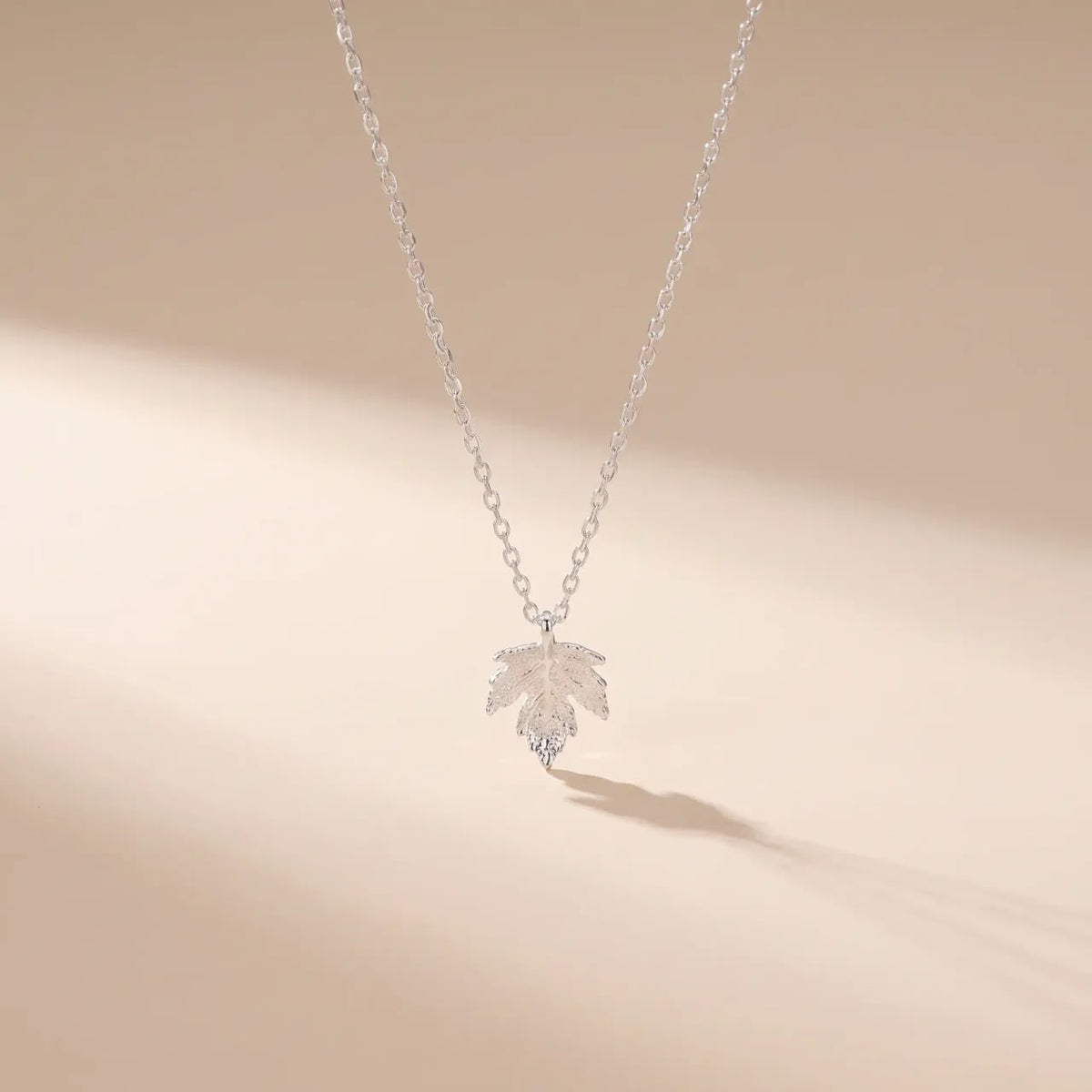 A delicate silver chain necklace with a small leaf-shaped pendant, photographed against a warm beige background with soft diagonal shadows.