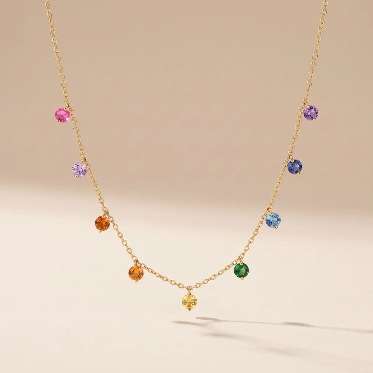 A delicate gold chain necklace featuring multiple small, colorful gemstone charms in pink, purple, blue, orange, yellow, and green, displayed against a soft beige background.