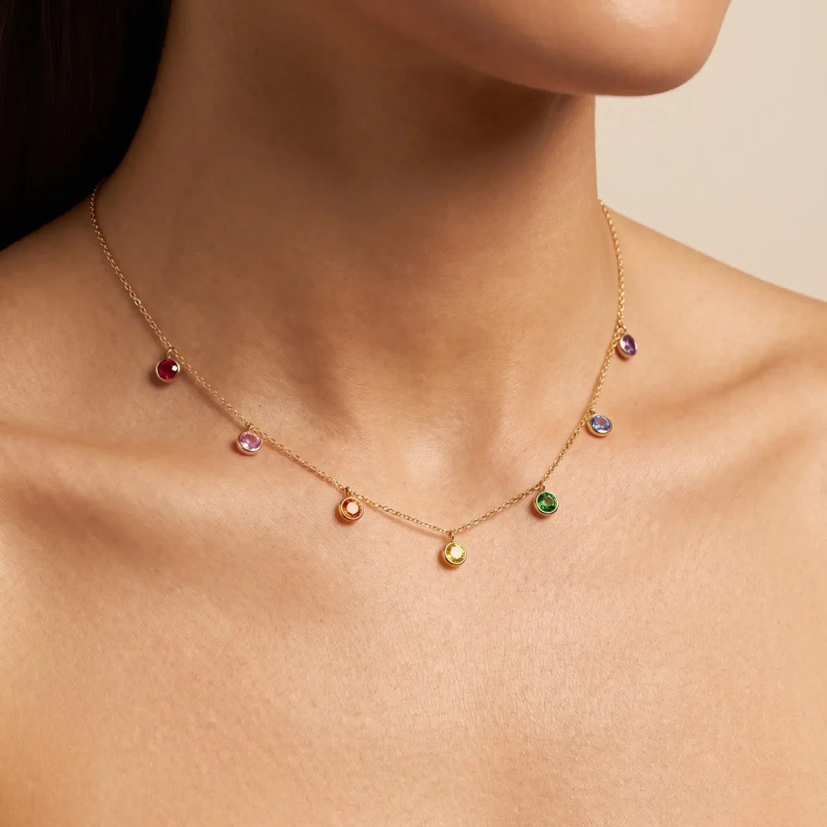 A delicate gold necklace with multiple small, colorful gemstone disc charms, shown worn on a model's neck against a soft beige background.