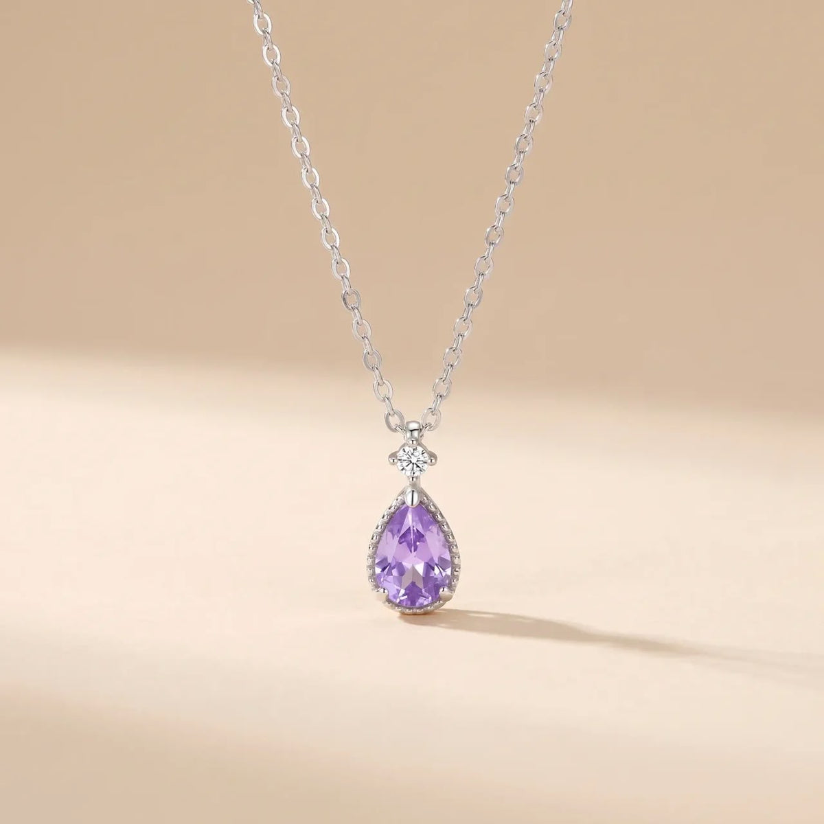 A silver necklace with a rectangular emerald-cut green gemstone pendant and small clear accent stones, shown against a warm beige background.
