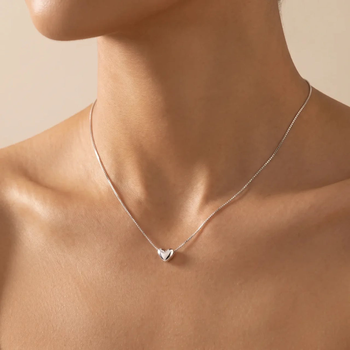 A close-up of a model's neck and upper chest wearing a delicate silver chain necklace with a small polished heart pendant.