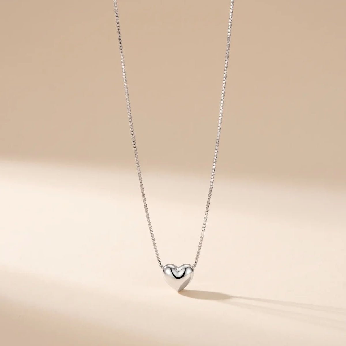 A delicate silver box chain necklace with a polished silver heart pendant, displayed against a warm beige background with soft shadows.