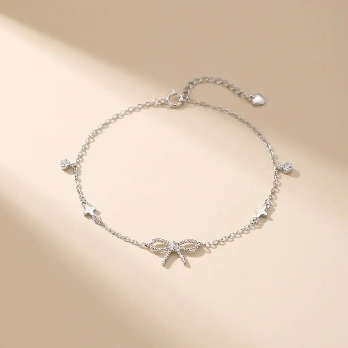 A delicate silver chain bracelet featuring a central bow charm, star accents, and small clear stones, displayed on a warm beige background with soft shadows.