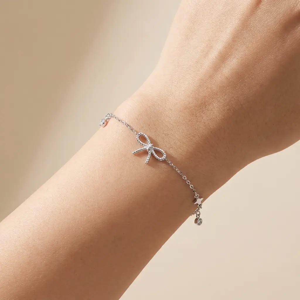 A delicate silver chain bracelet with a small bow charm and tiny dangling charms, shown worn on a person's wrist against a soft beige background.