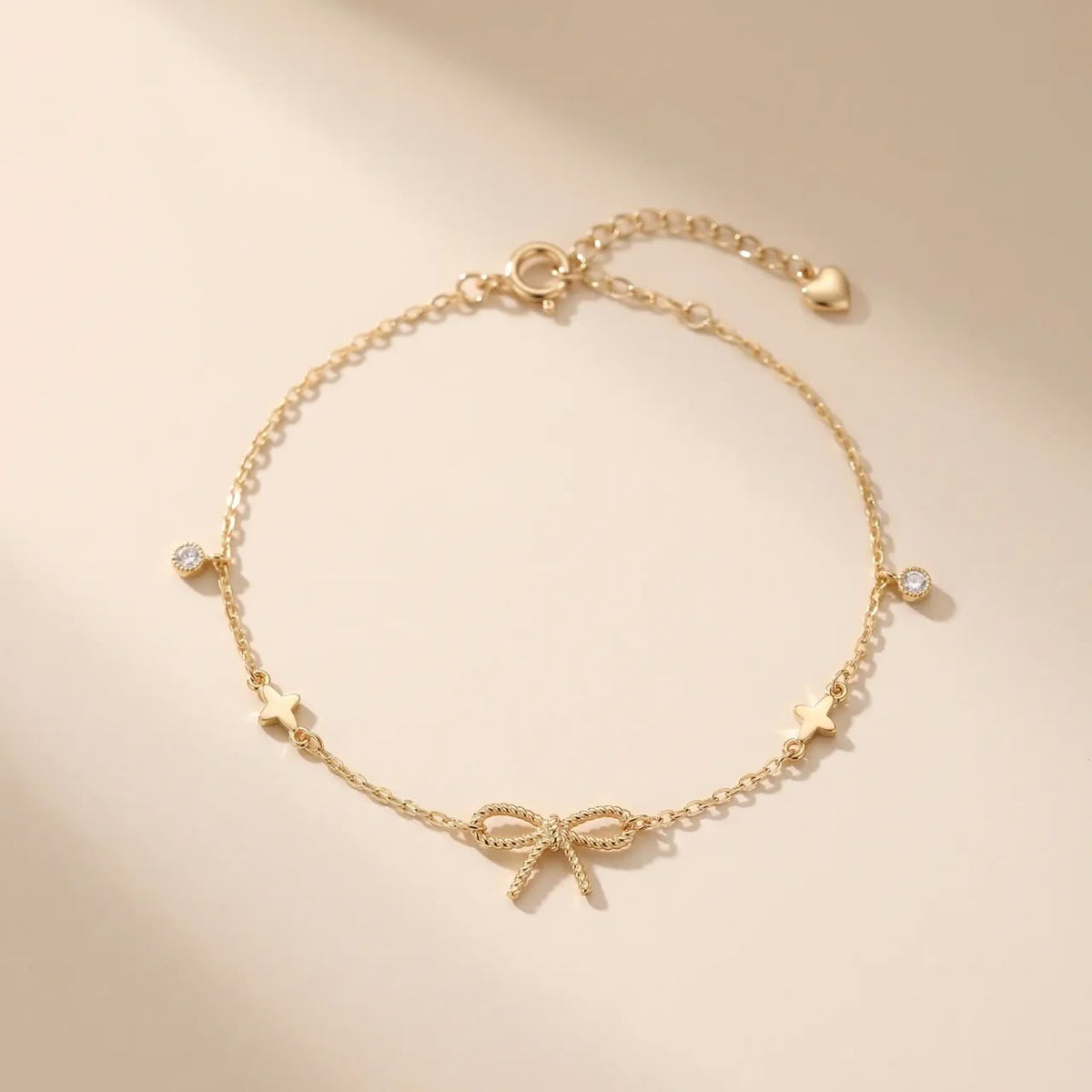 A delicate gold chain bracelet featuring a central textured bow charm, small star charms, and clear crystal accents, displayed on a warm beige background.