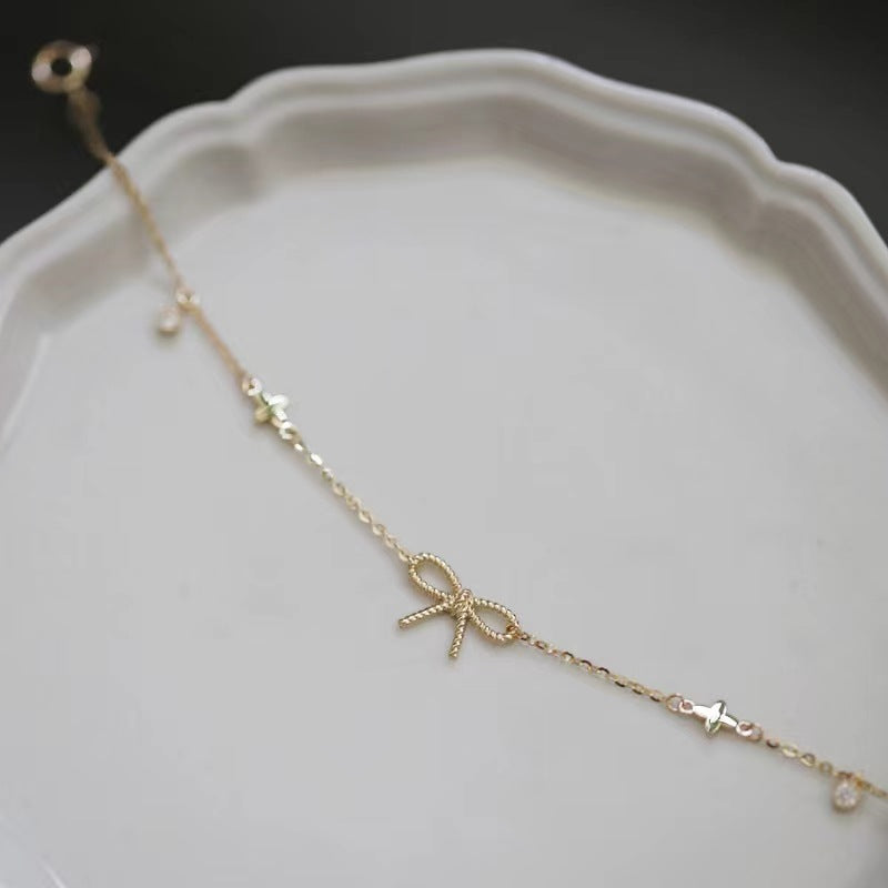 A delicate gold chain bracelet featuring a central bow charm, small star details, and tiny clear stones, displayed on a white ceramic dish.