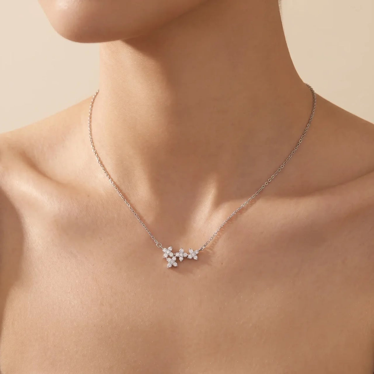 A delicate silver chain necklace with a small cluster of white flower-shaped charms, shown worn on a model's neck against a warm beige background.