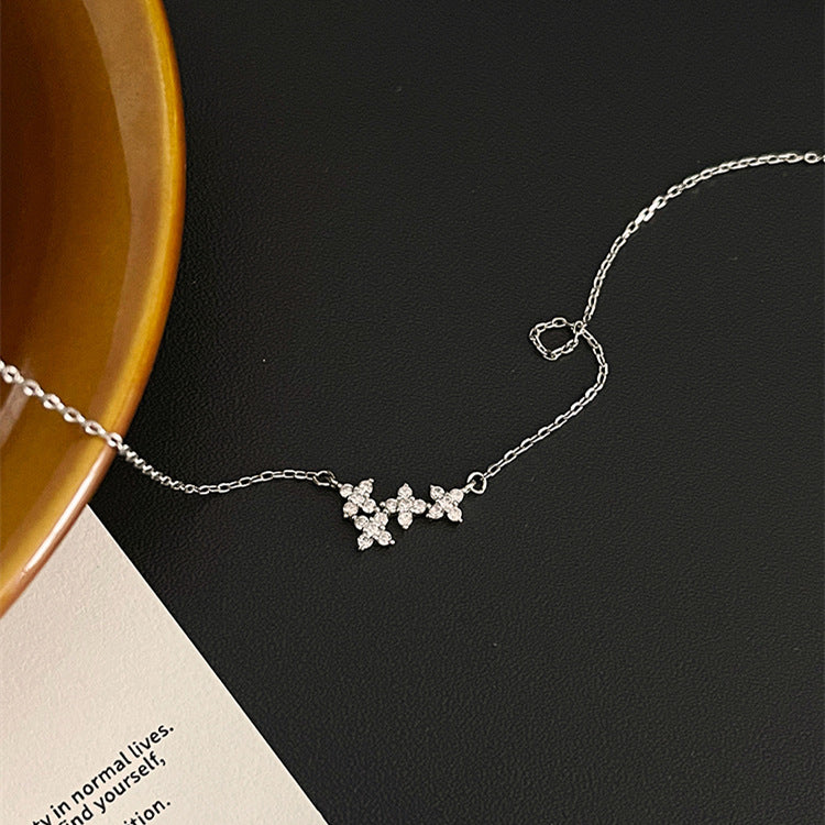 A delicate silver chain necklace with three small flower-shaped crystal clusters, styled on a dark textured surface next to a brown ceramic bowl and a corner of a printed card.