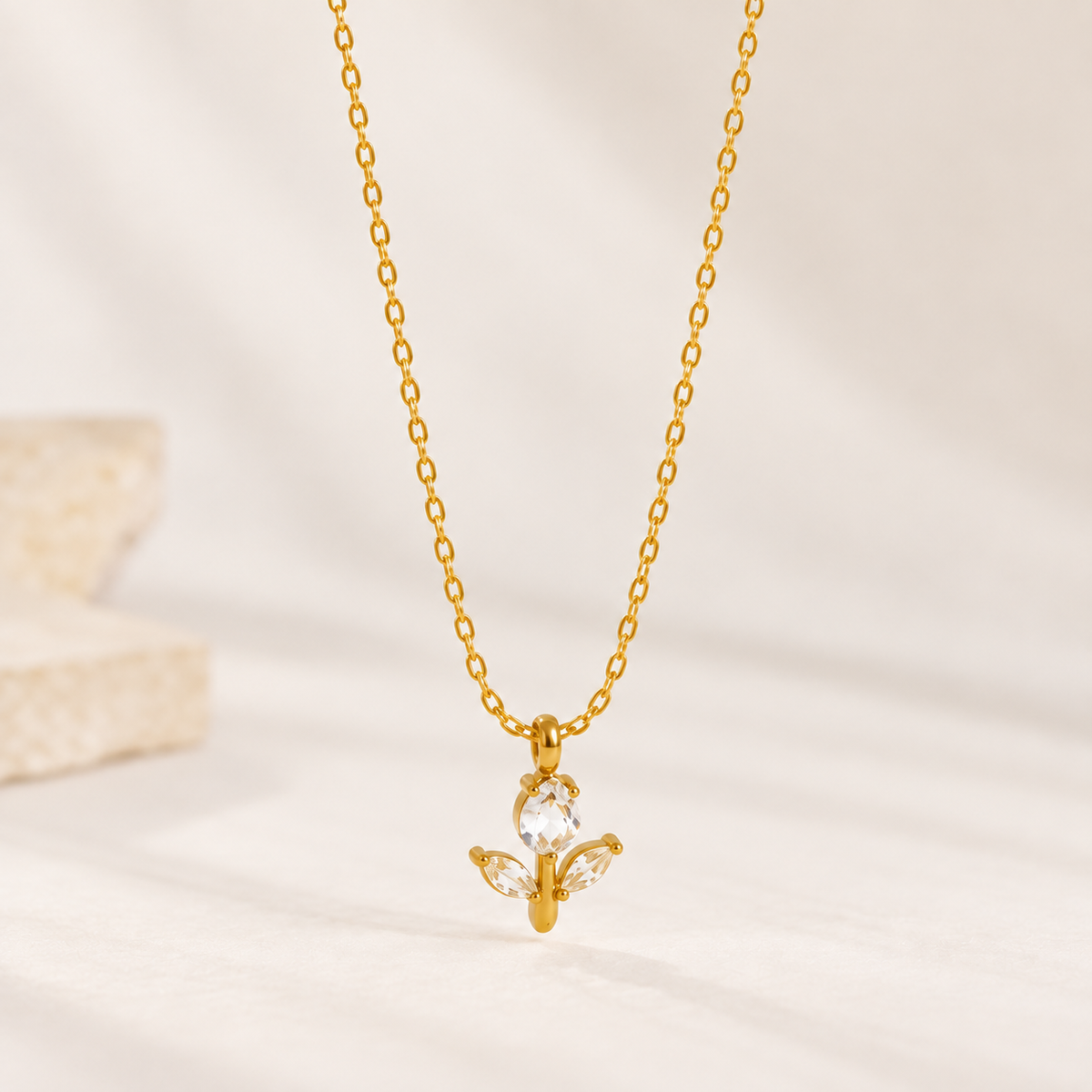 A delicate gold chain necklace with a small pendant featuring a clear faceted gemstone and three small clear crystal leaves, displayed against a soft, light beige background.