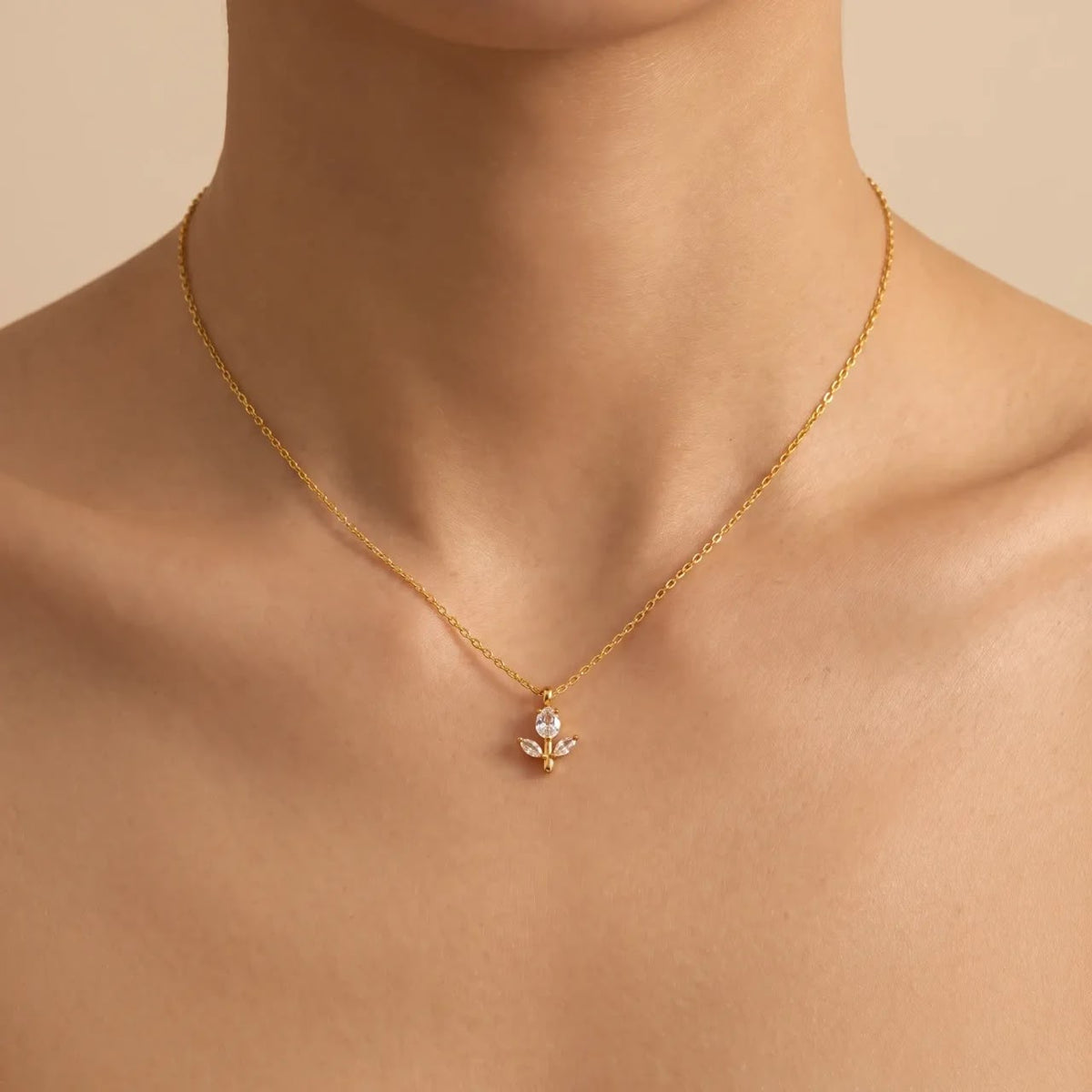 A delicate gold chain necklace with a small floral pendant featuring clear stones, shown worn on a model's neck against a warm beige background.