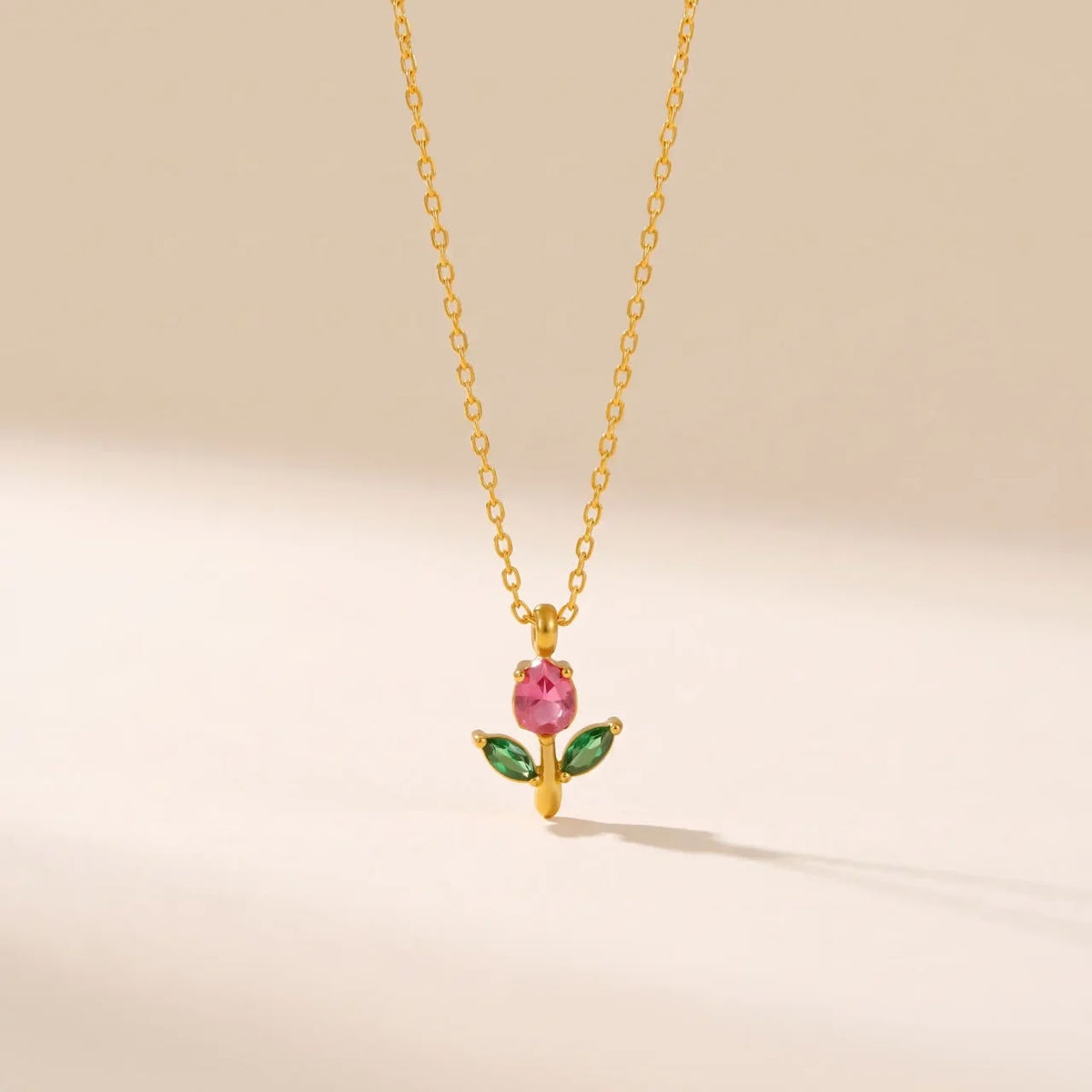 A delicate gold chain necklace featuring a small pendant shaped like a flower, with a pink gemstone center and two green marquise-cut stones on the sides, set against a warm beige background.