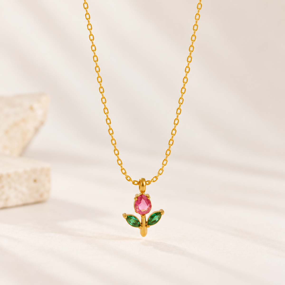 A delicate gold chain necklace with a small pendant shaped like a flower, featuring a pink gemstone center and two green gemstone leaves, displayed on a soft beige fabric background.