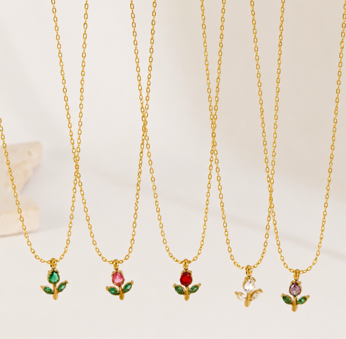 Five gold-tone floral pendant necklaces with small flower charms are arranged in a fan shape against a light cream background. The pendants feature green leaves, a pink flower, a red flower, a clear gem, and a purple gem.