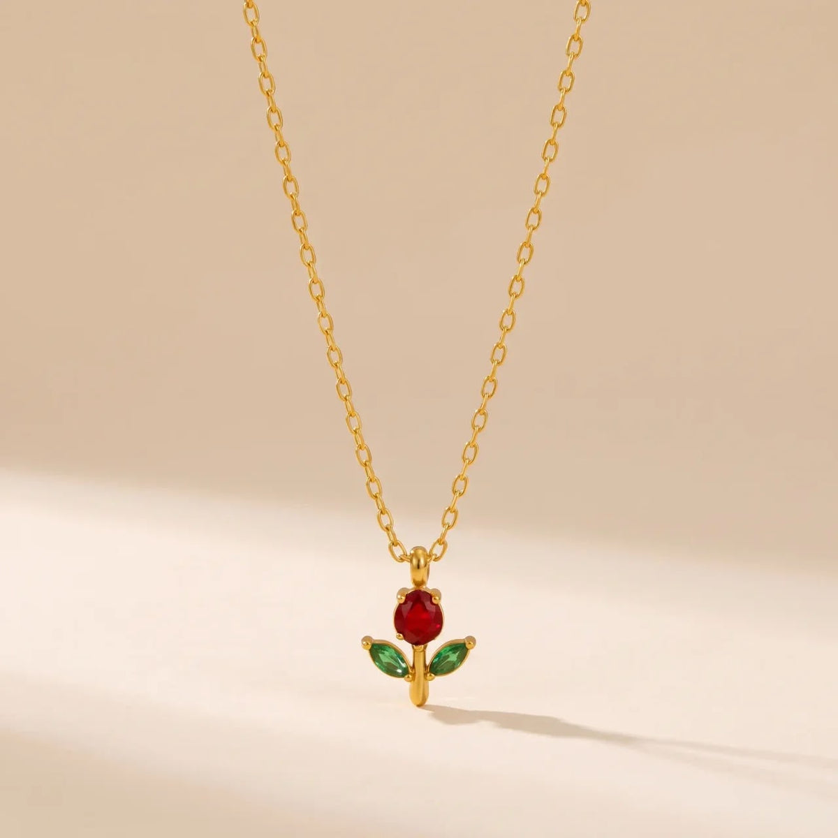 A delicate gold chain necklace featuring a pendant with a red rose bloom and green leaves, displayed against a warm beige background.