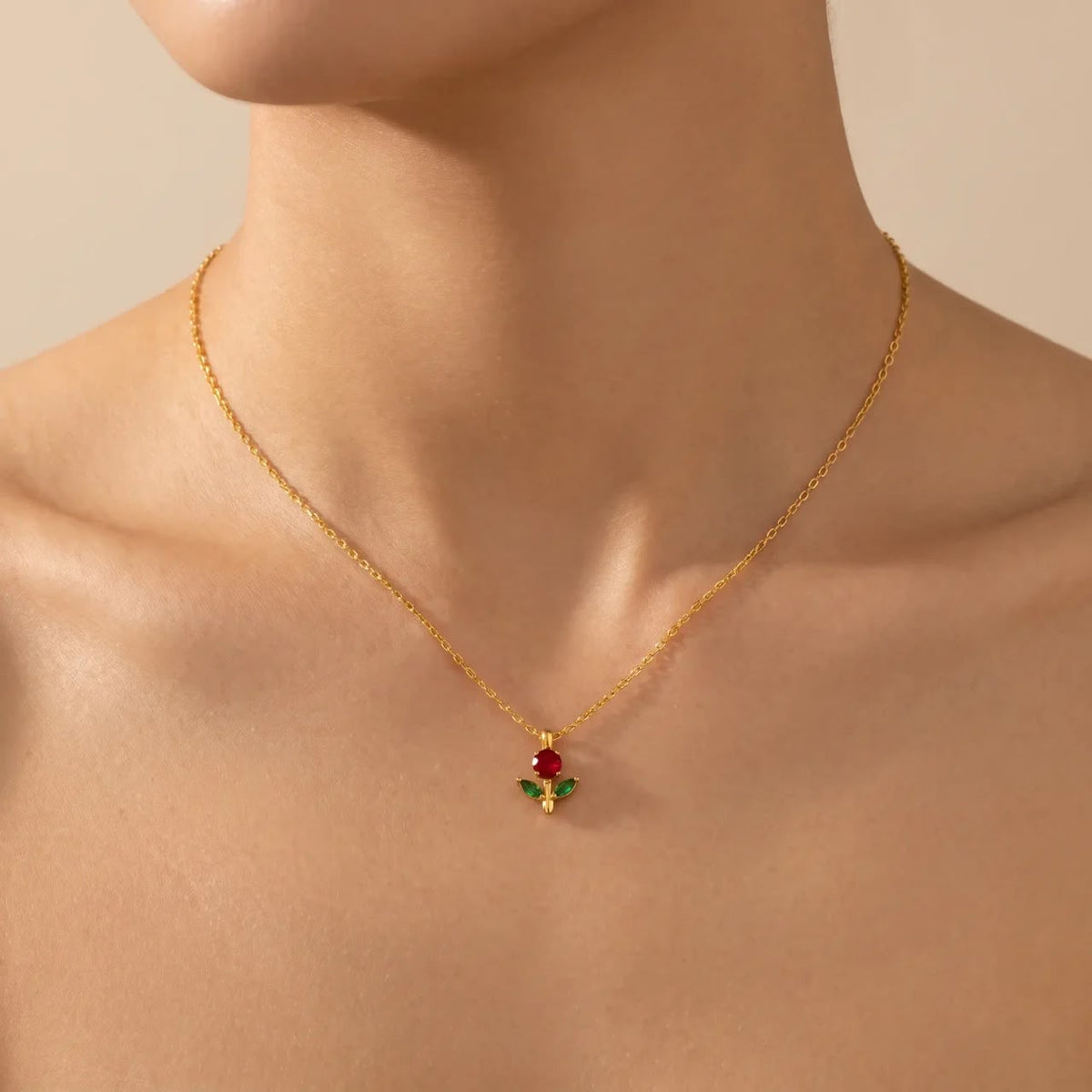 A delicate gold chain necklace with a small pendant featuring a red rose and green leaves, shown worn on a model's neck against a warm beige background.