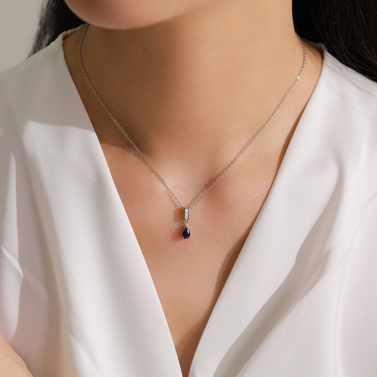 A close-up of a model wearing a delicate silver chain necklace with a teardrop-shaped dark blue pendant and a small clear stone accent.