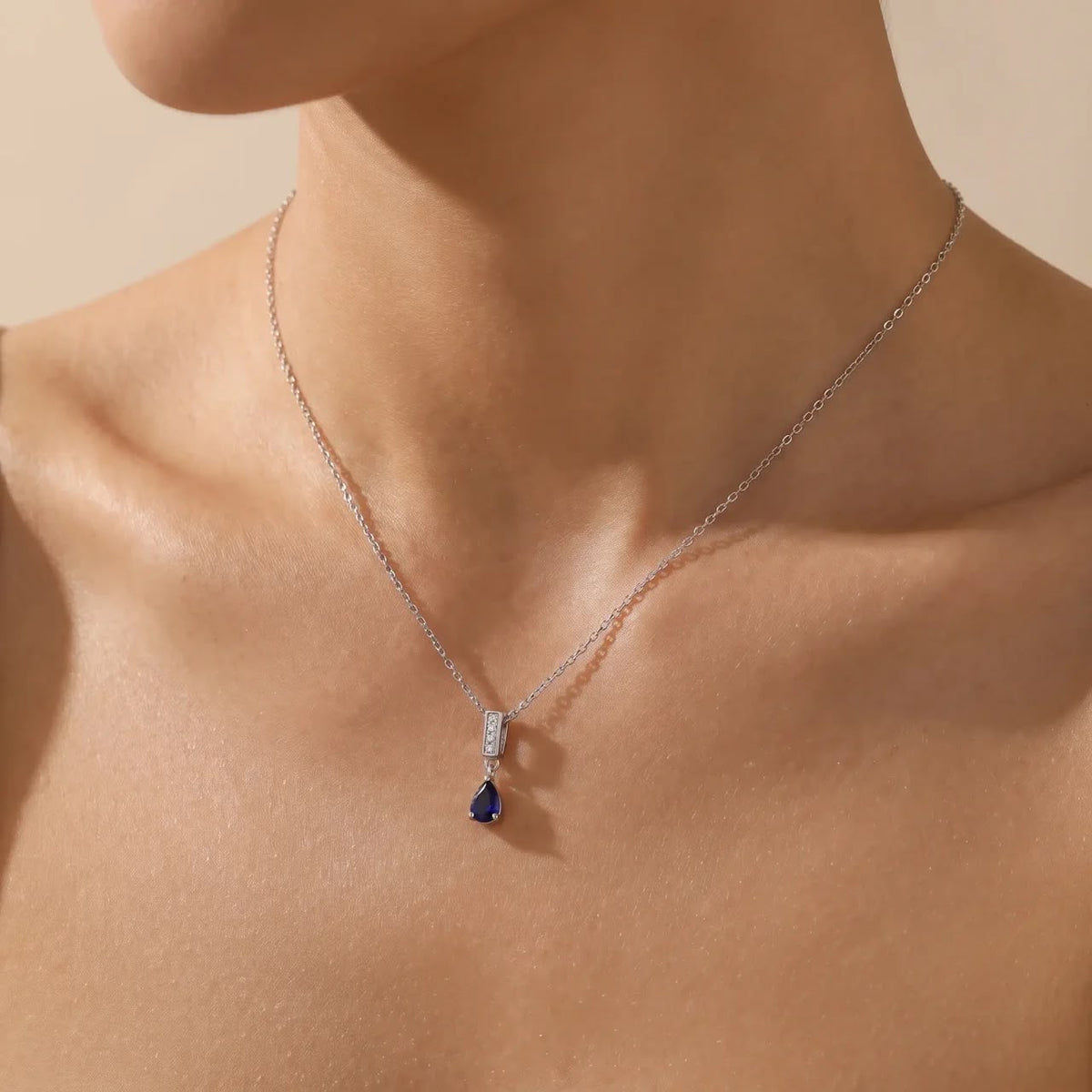 A close-up of a model's neck and upper chest wearing a delicate silver chain necklace with a pear-shaped dark blue gemstone pendant.
