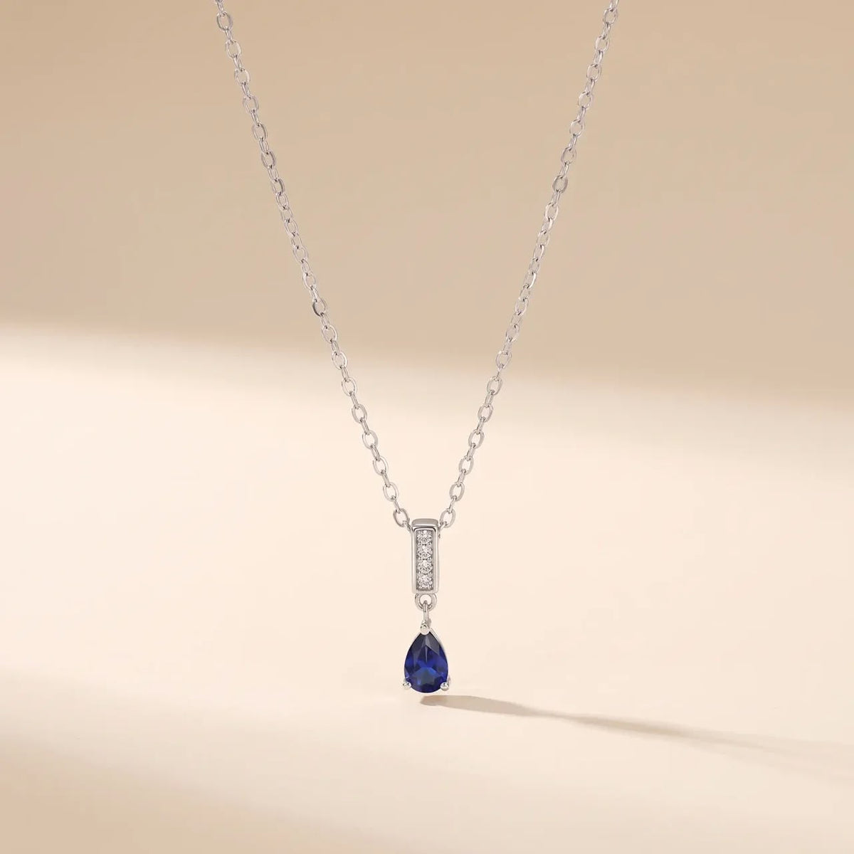 A delicate silver chain necklace featuring a pear-cut dark blue gemstone pendant, accented by a vertical bar of small clear stones, set against a soft beige background.