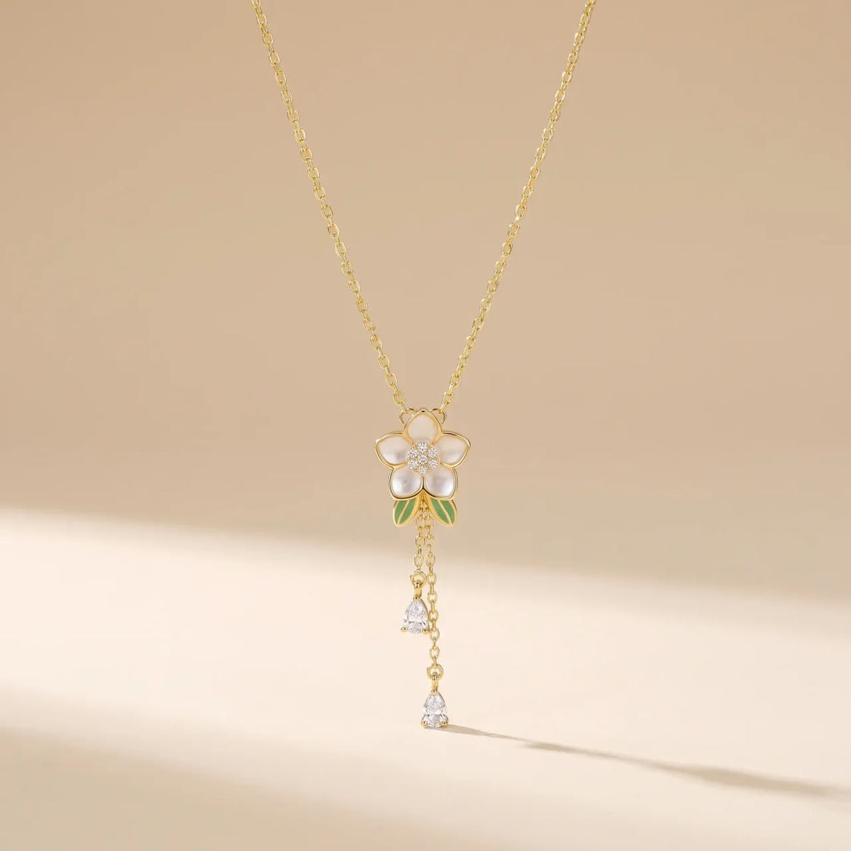 A delicate gold chain necklace featuring a flower pendant with white mother-of-pearl petals, green enamel accents, and dangling clear crystal charms, set against a warm beige background.
