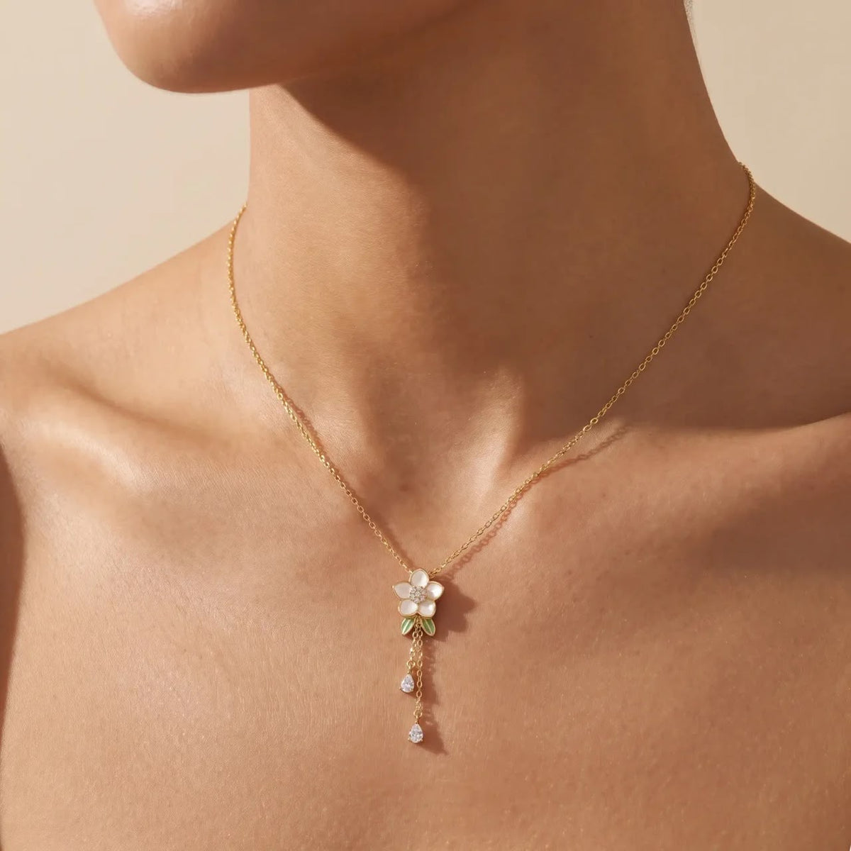 A delicate gold chain necklace with a small white flower pendant and dangling pearl-like drops, shown worn on a model's neck against a warm beige background.