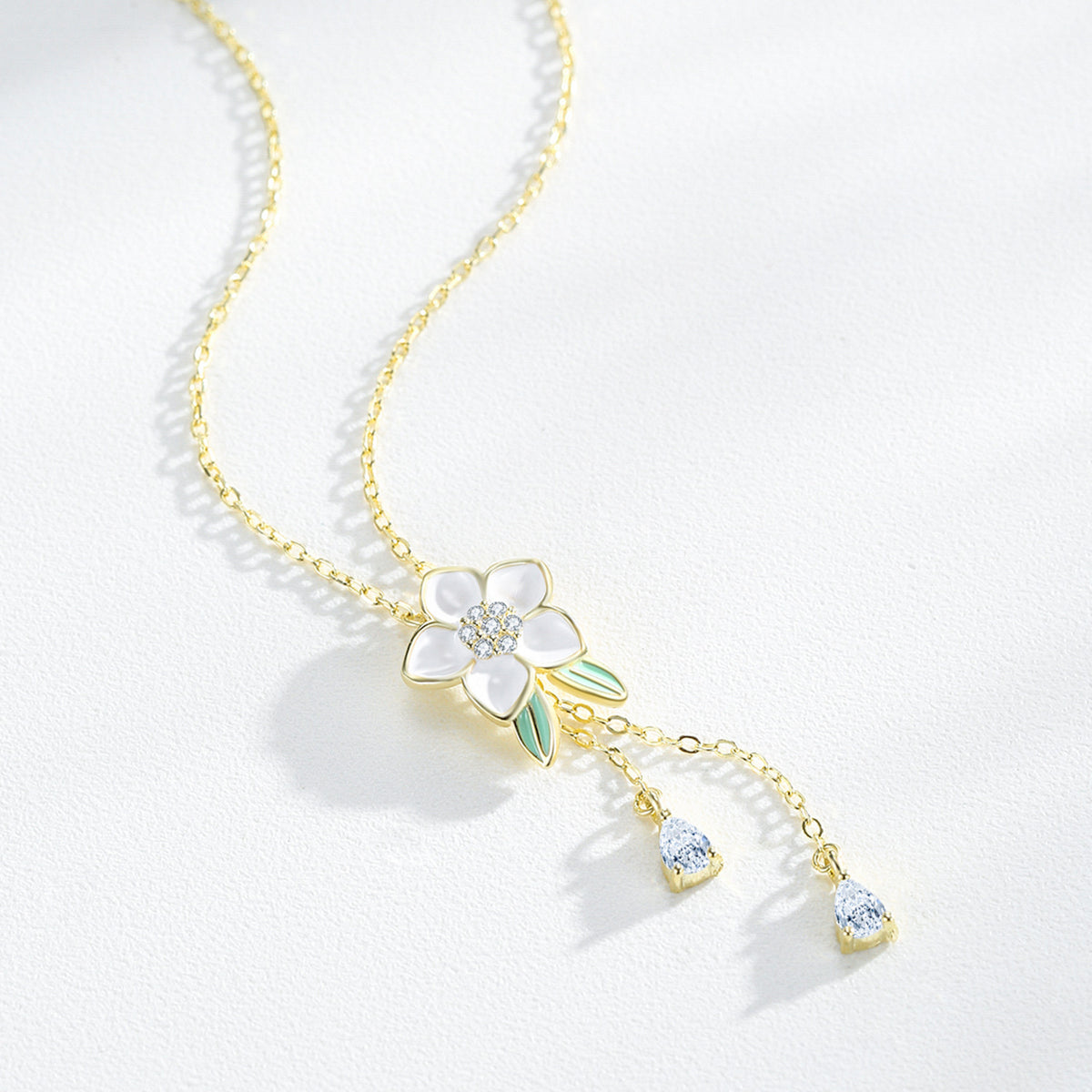 A delicate gold chain necklace featuring a flower pendant with white enamel petals, a small clear crystal center, and light green enamel leaves. Two small clear crystal teardrop charms dangle from the bottom of the flower on a white textured background.